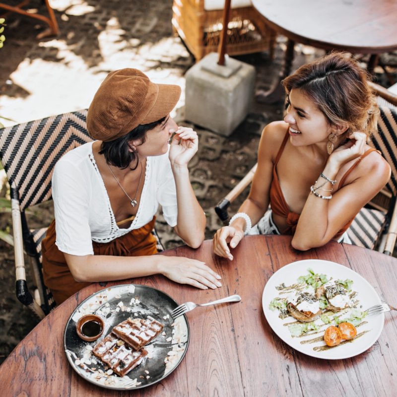 Brunchs D'Automne À Pornichet : Adresses Cosy Et Cuisine Réconfortante Au Bord De L'Eau 3 View From Above At Tanned Girls In Stylish Outfits Talking And Enjoying Tasty Food In Street Cafe. Pretty Woman In Cap And Lady In Brown Bra Smile And Eat Waffle Outside